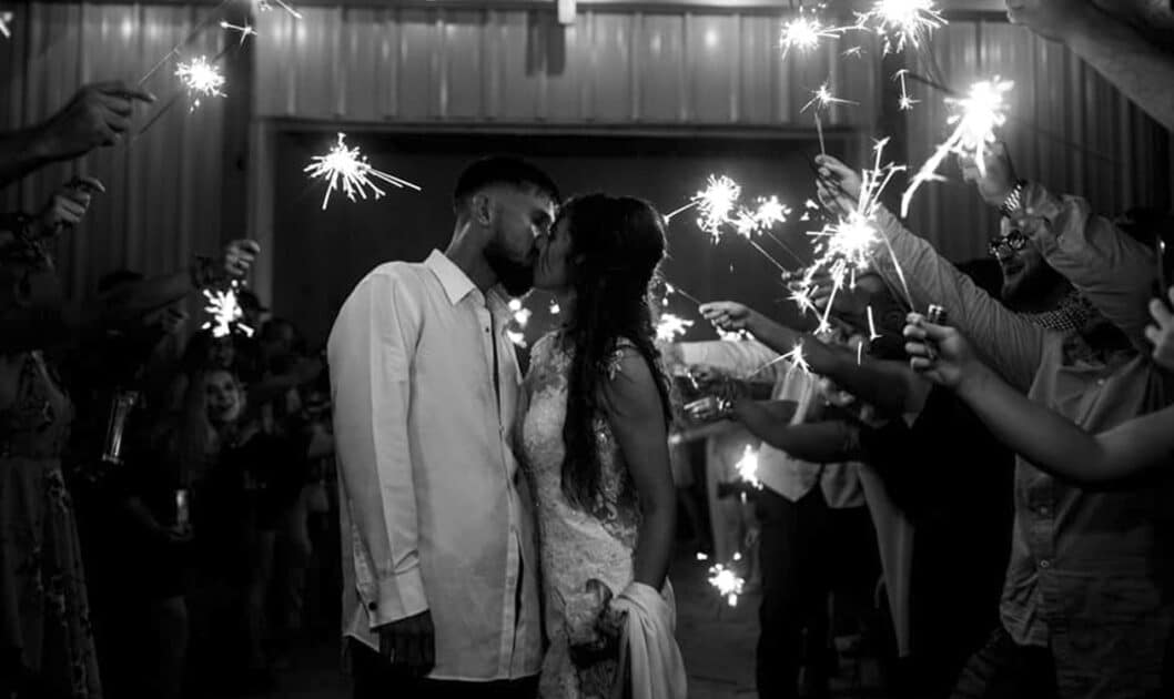 A black and white photo of a bride and groom sharing a kiss surrounded by their friends holding sparklers
